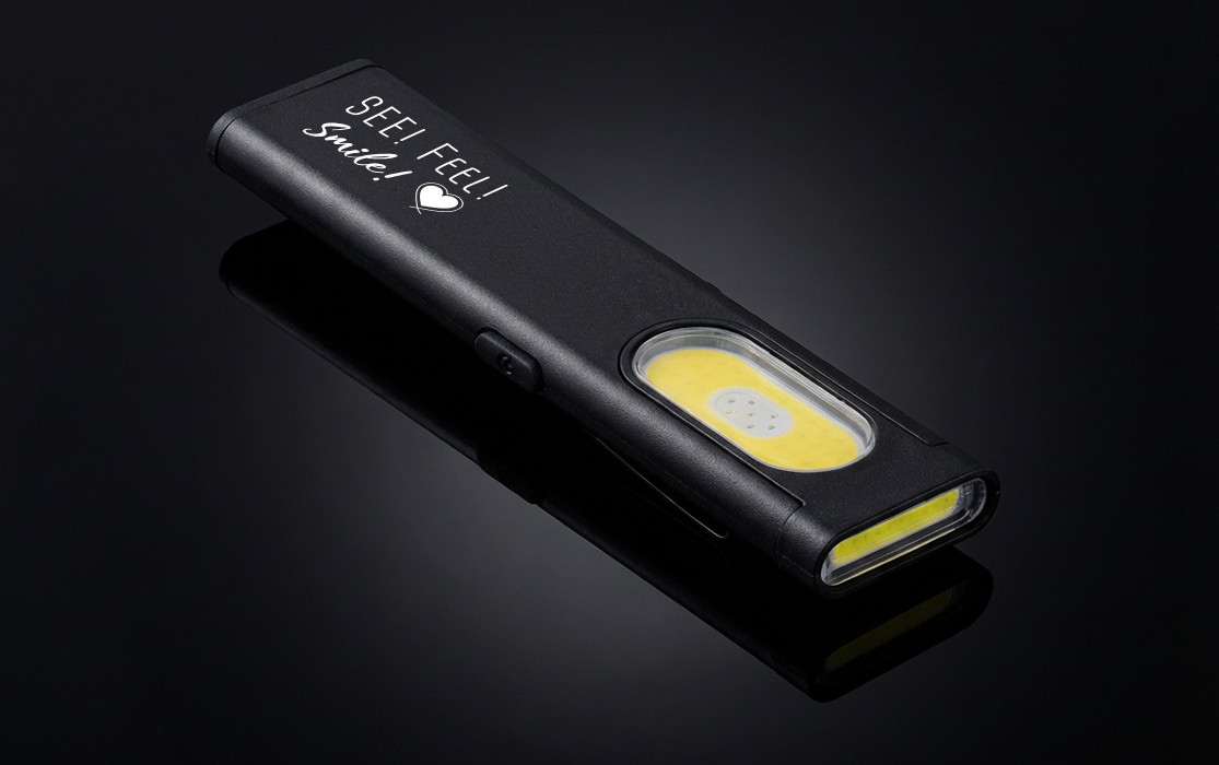 Black, compact work light with engraving "SEE! FEEL! Smile!" on a black background