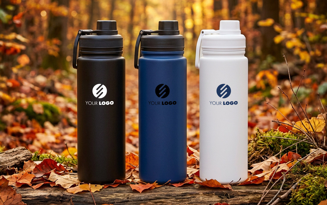 Drei ARCTICDROP Trinkflaschen in Schwarz, Blau und Weiß mit Logofläche, auf einem Waldboden mit buntem Herbstlaub.