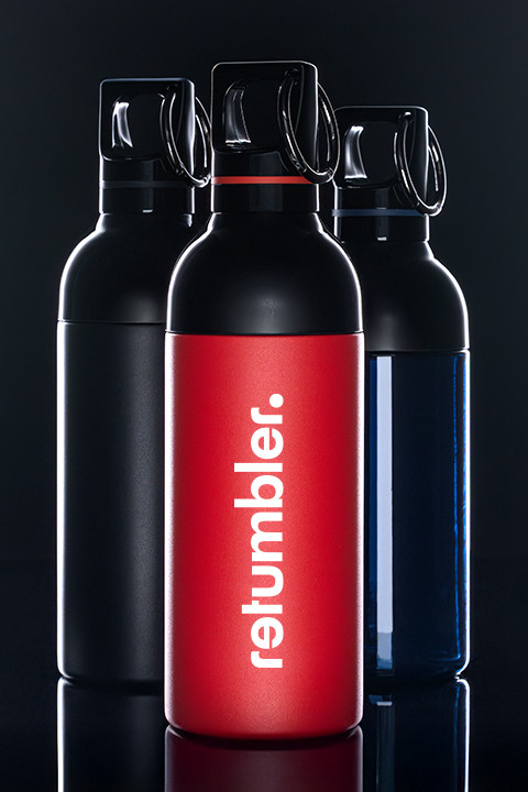 Drie retumbler roestvrijstalen drinkflessen in zwart, rood en blauw, met de rode fles en verticaal retumbler‑logo op de voorgrond – duurzame herbruikbare drinkware.