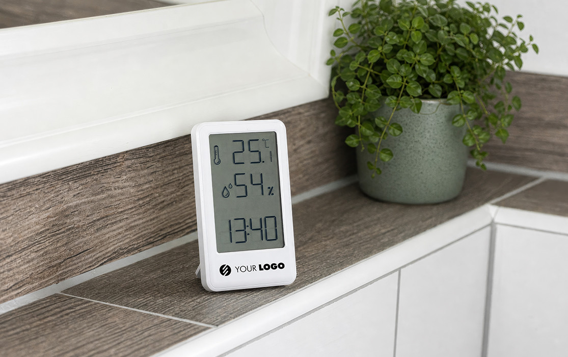 Digital thermo‑hygrometer with logo space Digital thermometer with hygrometer and clock, featuring a branding area for your logo, on a windowsill next to a houseplant