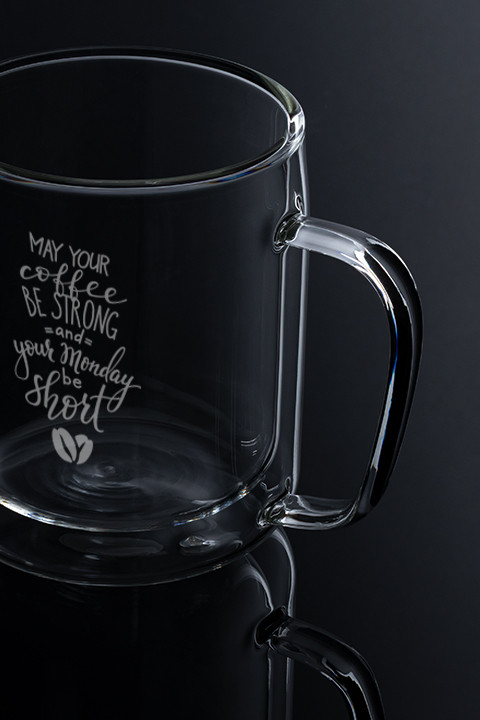 Glasbecher mit Spruchgravur Transparente Glastasse mit graviertem Spruch „May your coffee be strong and your Monday be short“ auf dunklem Hintergrund.