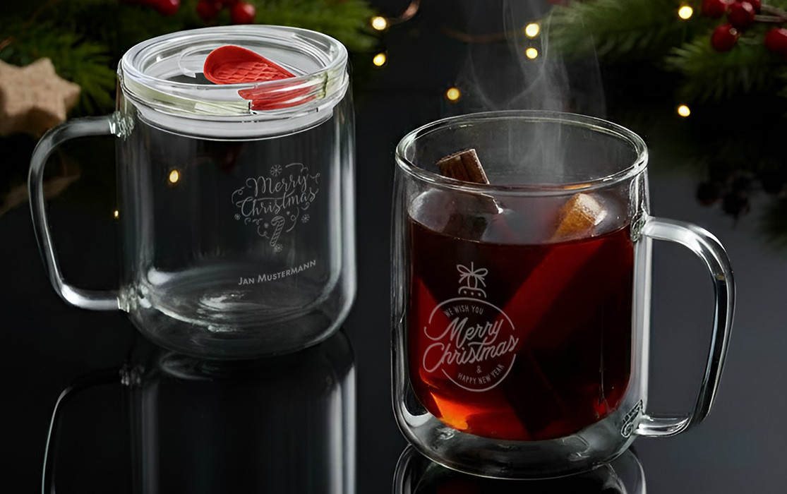 Zwei transparente Glasbecher mit Weihnachtsmotiv, einer mit heißem Getränk und Zimtstange, vor weihnachtlicher Dekoration.