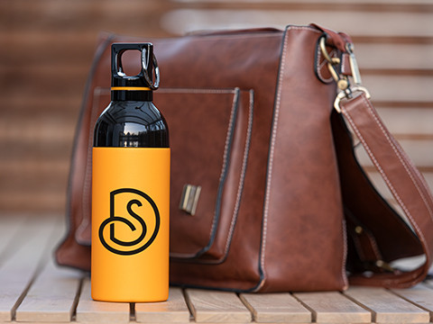 Gele retumbler roestvrijstalen drinkfles met zwart logo, geplaatst voor een bruine leren tas – perfect voor dagelijks gebruik.