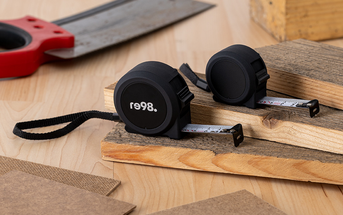 Two black RE98-TAPEA measuring tapes for 3 meters and 5 meters on wood, tools in the background
